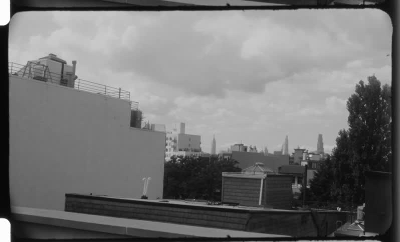 A black and white view of city buildings and rooftops with trees showing distant skylines under a cloudy sky framed by the edges of 16mm film stock