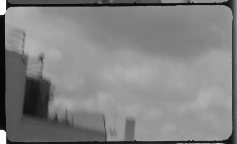 A black and white film frame shows a cloudy sky over dense urban rooftops with distant city skyscrapers visible