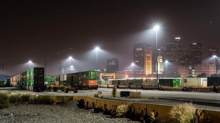 A long exposure time lapse shot at night shows trains passing through an illuminated intermodal freight yard full of green shipping containers with the bright skyline of a large city in the background