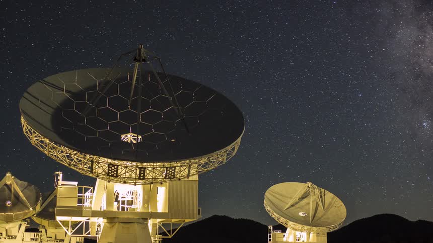 A time lapse shot shows large radio telescope dishes rotating under a dark star filled night sky with the faint visible streak of the milky way