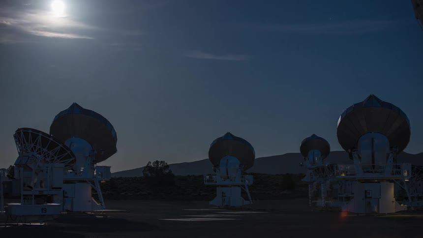 A field of large radio telescope dishes stands under a dark blue night sky with a bright moon