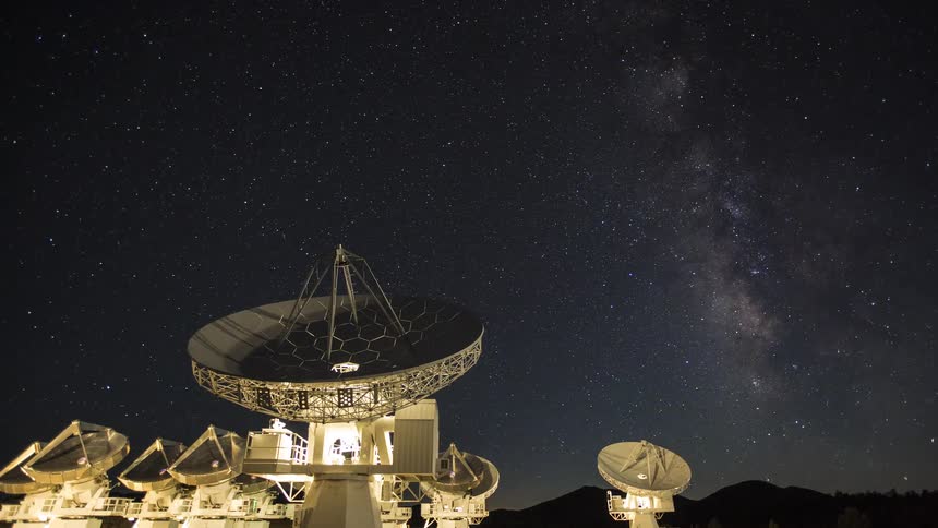 A long exposure time lapse shot at night shows a large array of white radio telescope dishes rotating slowly beneath a dark sky filled with bright stars and the distinct milky way galaxy