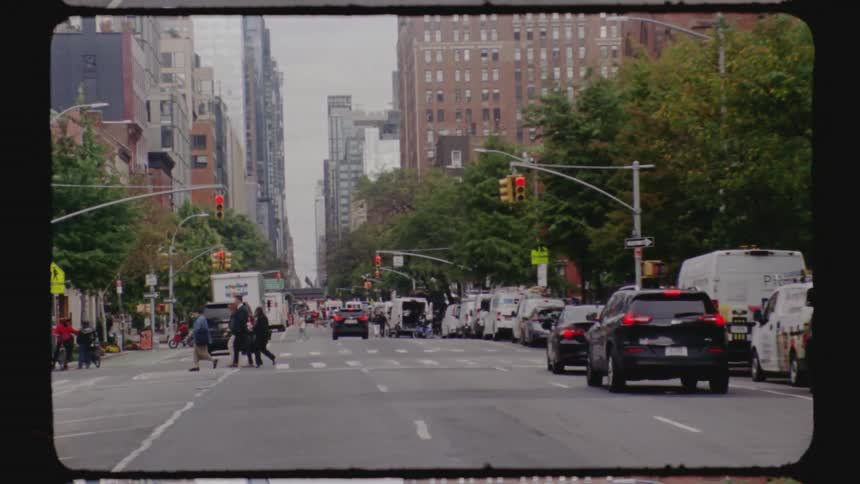 Pedestrians crossing a wide avenue with heavy traffic between tall buildings and treelined areas