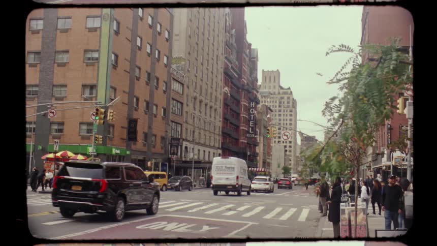 A vintage film style shot captures a busy street corner in new york city with tall apartment buildings and a red brick hotel with a large black vertical sign for the chelsea hotel