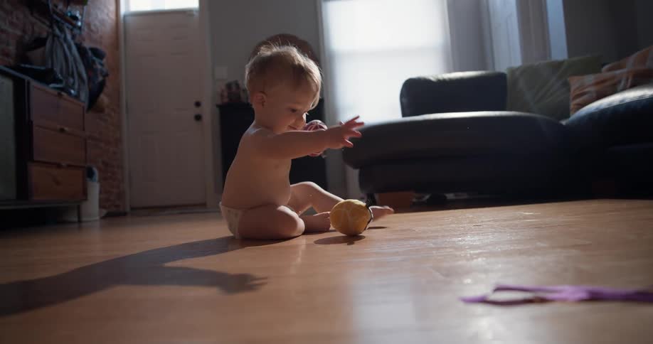 A happy baby in a diaper plays on a sunlit wooden floor in a living room reaching for a small ball then picking up two patterned balls and holding them in their hands before looking toward the camera