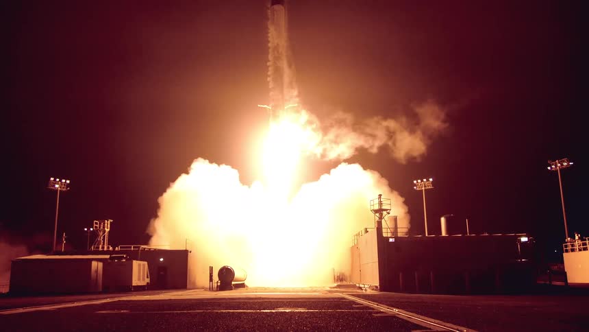 A sleek rocket erupts upward at night its engines blazing with fire and smoke as it ascends from a brightly lit launch pad