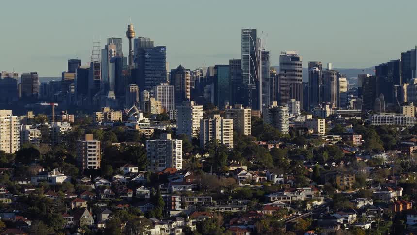 A large citys dense metropolitan skyline featuring modern glass towers and highrise apartment blocks rising above a foreground of lush treefilled suburban houses