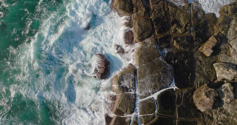Waves crashing against geometric rock formations seen from above