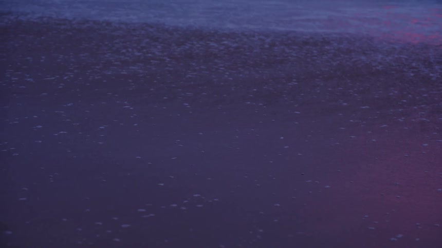 A closeup view captures the tide gently washing over the sandy shore with the shallow water and foam reflecting deep purple and pink hues from the twilight sky