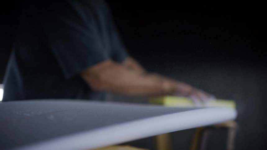 A closeup shot focuses on a persons hands rhythmically sanding the smooth white surface of a surfboard with a yellow block creating fine dust particles that float in the air