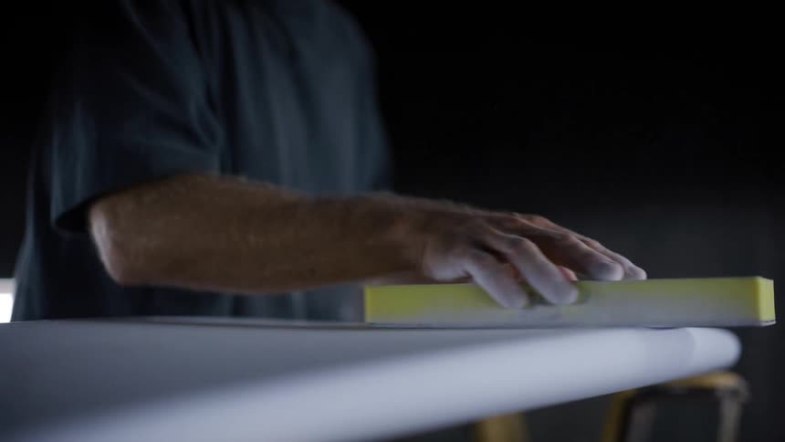 A person sands the rail of a white surfboard using a yellow sanding block