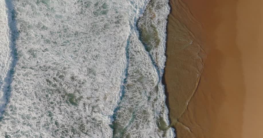 An overhead static drone shot captures the mesmerizing action of ocean waves continually crashing and receding against a line of wet goldenbrown sand forming a thick layer of white foam