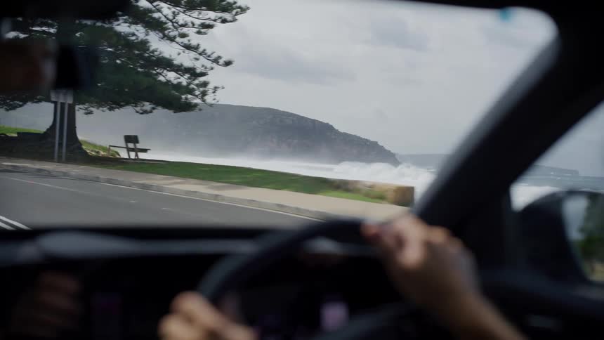Driver steering the car along a coastal road with a rocky peninsula in the background