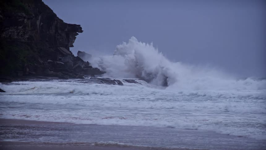 A wide shot captures powerful ocean waves crashing dramatically against dark coastal rocks and cliffs under a heavy overcast blue sky