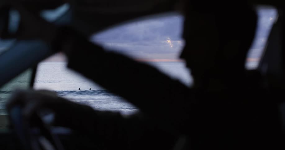 Man getting into the car with a view of surfers in the ocean through the open window