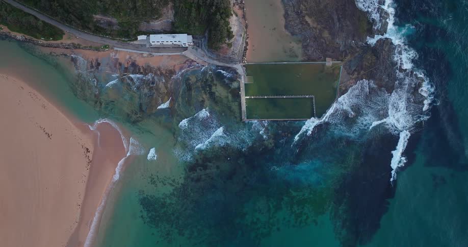An overhead drone shot captures a rectangular green ocean fed tidal pool next to a sandy beach and rocky coastline with crashing turquoise waves