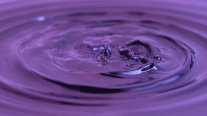 A closeup shot captures a water droplet hitting a pool of purple liquid creating a concentric ripple pattern that gives way to a complex beautiful and turbulent crown splash