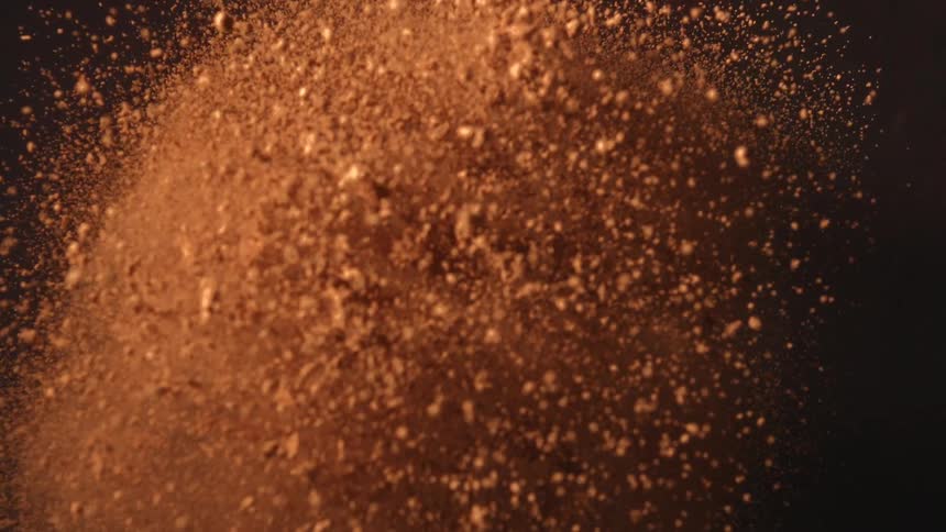A large cloud of fine reddish brown powder explodes outward and slowly settles against a dark background in a dynamic burst of movement