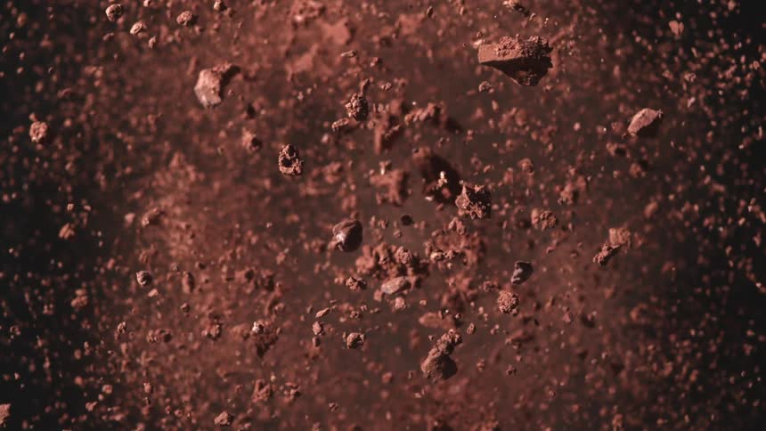 A large mound of brown clumpy soil or powdered material rapidly expands and explodes upward and outward into thousands of suspended particles against a black background in slow motion