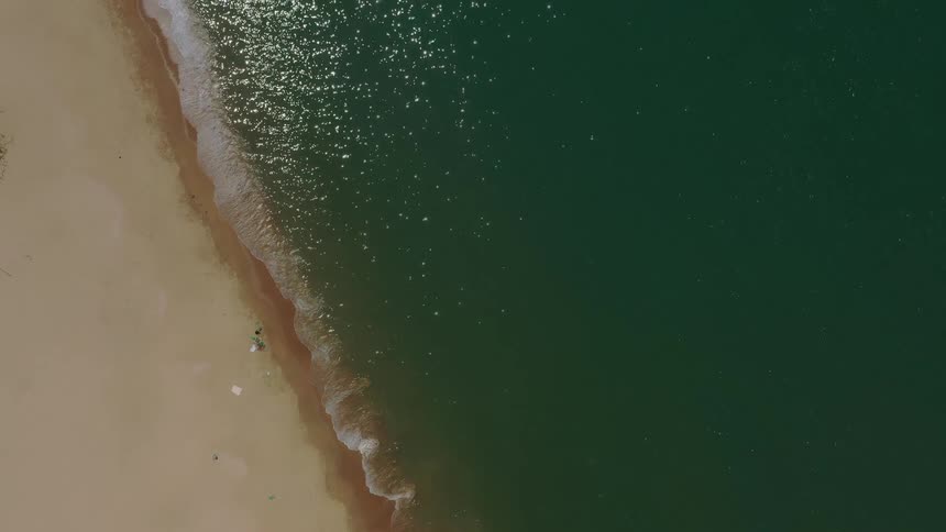 An overhead drone shot captures the dynamic border between a stretch of beige sand and the deep green ocean water as small white waves consistently break along the shoreline