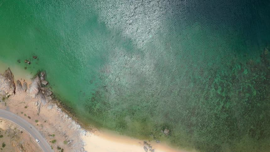 An extreme overhead shot captures the distinct contrast between the shallow clear turquoise ocean and the deeper water bordered by a beige sandy beach and a curved coastal road along a rocky bank