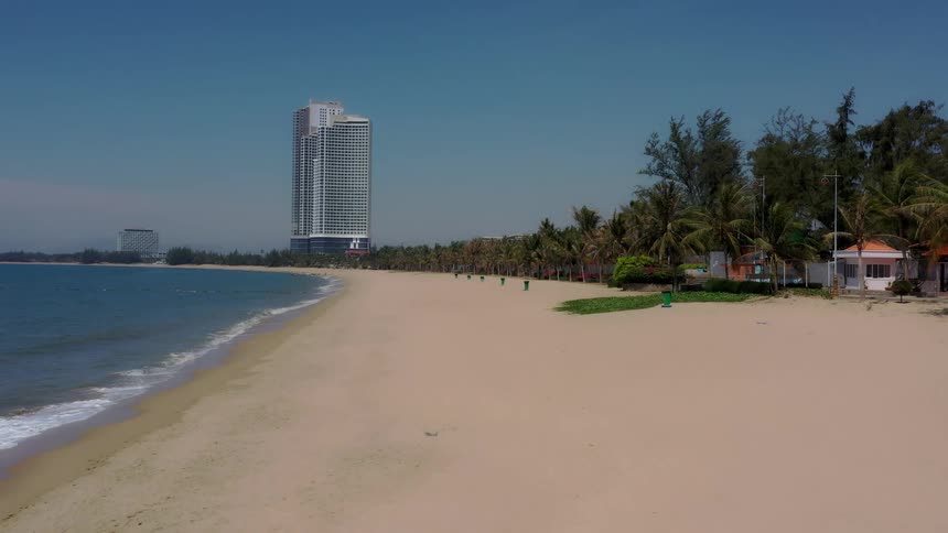 A long empty sandy beach stretching towards the calm ocean with a tall modern white skyscraper dominating the background behind a row of palm trees and small coastal structures