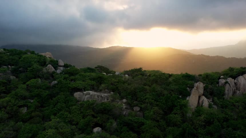 Sunbeams break through dark clouds over a brushcovered mountain landscape with many prominent rocks at sunset