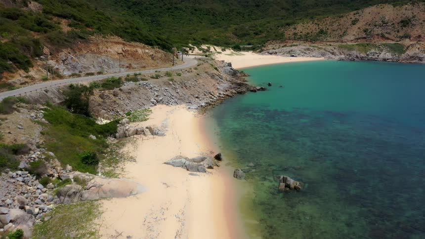 An aerial drone shot slowly flies over a sandy secluded cove revealing a winding paved road hugging the edge of the steep greencovered hillside that meets the strikingly clear turquoisecolored water