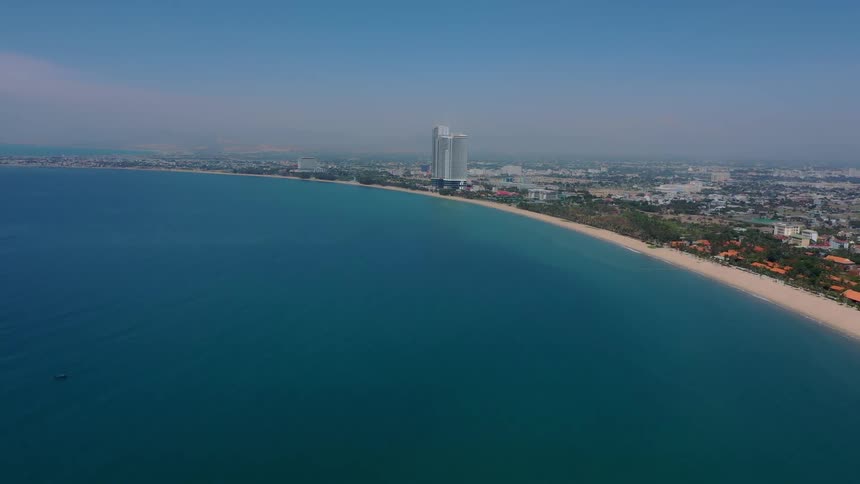 A tall modern building and a city stands on a long sandy beach next to a vast blue ocean