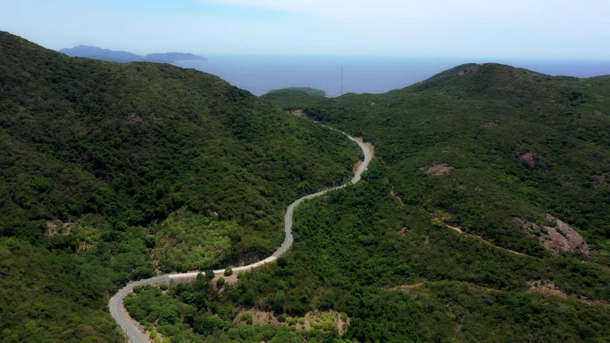 A winding grey road snakes through steep thickly forested green mountains