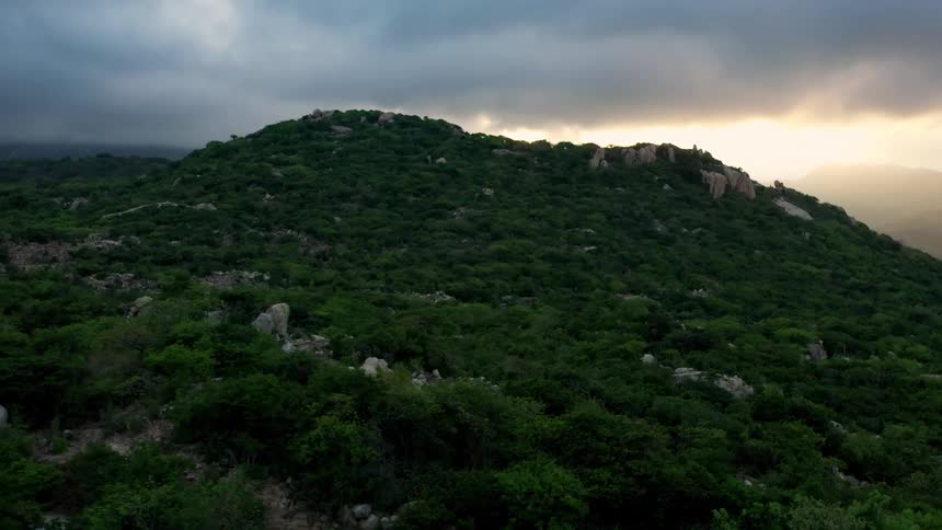 A thickly vegetated hill rises under a dark cloudy sky with a bright patch of sunlight on the right