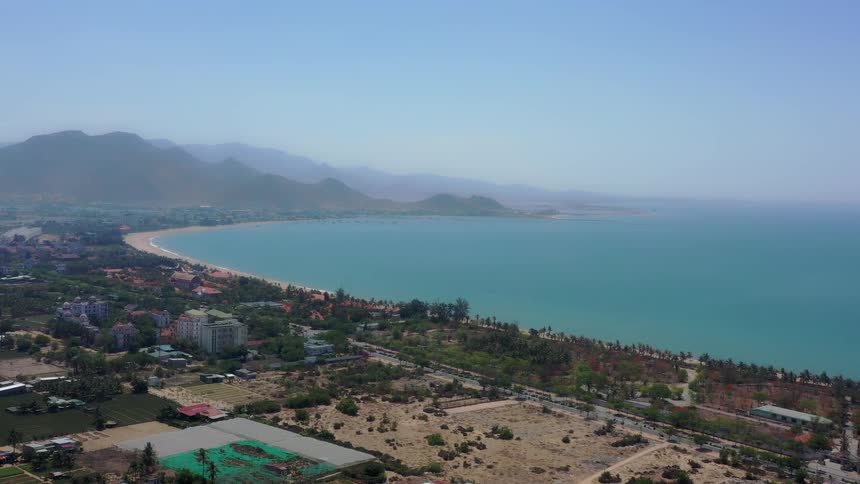 An expansive aerial shot slowly descends over a tropical coastal town showcasing houses and farmland leading to a long crescentshaped sandy beach bordered by deep blue ocean and framed by distant hazy mountains