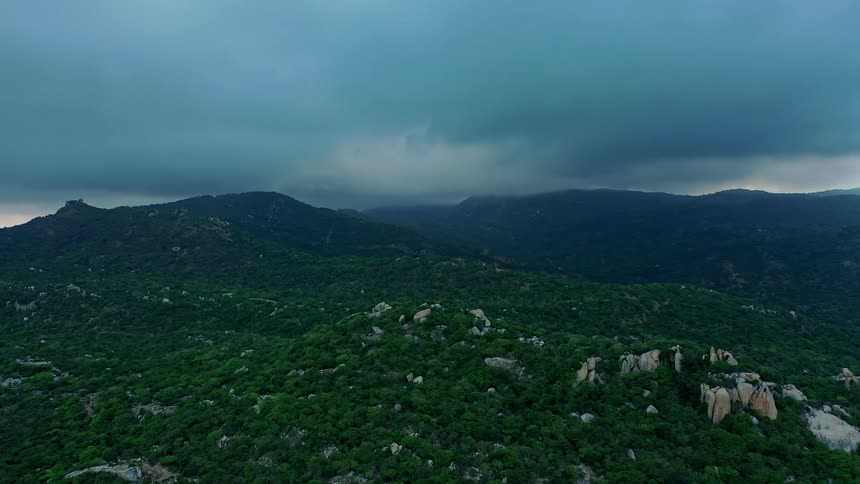 An aerial view slowly drifts over a rugged landscape of rolling hills covered in dense green foliage and dotted with large rocks all shrouded by a dramatic and dark heavy layer of overcast clouds