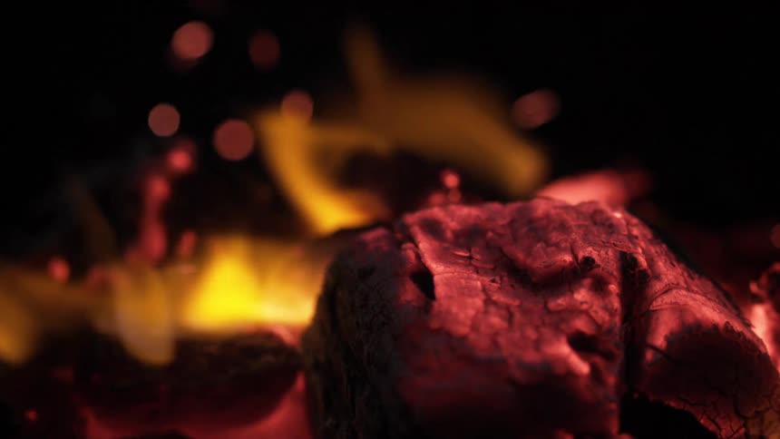 A dramatic closeup focuses on a piece of bright red glowing charcoal as brilliant yellow and orange flames surge around it casting heat and sending small sparks into the dark background