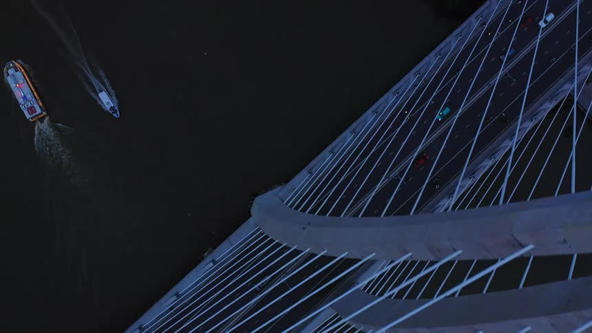 An overhead drone shot captures traffic moving across a modern illuminated cablestayed bridge over dark water while a few small boats move diagonally underneath it