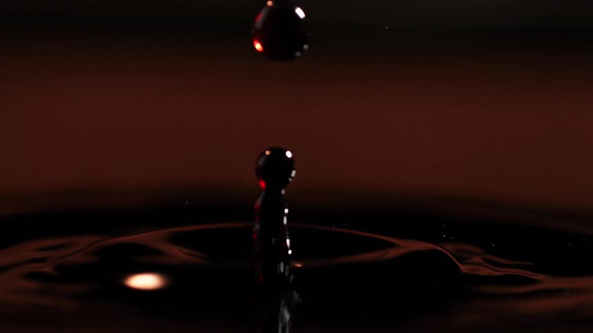 A droplet of water falls into a glass of red wine creating ripples that spread across the surface and distort the deep crimson liquid with gentle waves