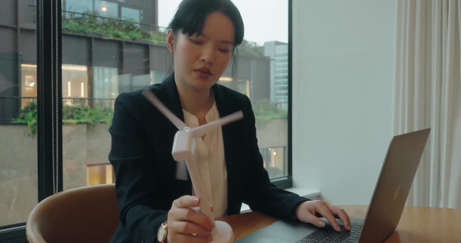 Woman working in an office observes a small wind turbine model before typing on a laptop