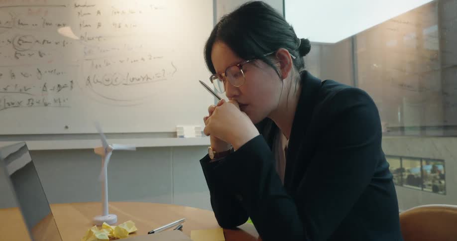 A businesswoman sits thoughtfully beside a laptop and a model wind turbine in a modern office