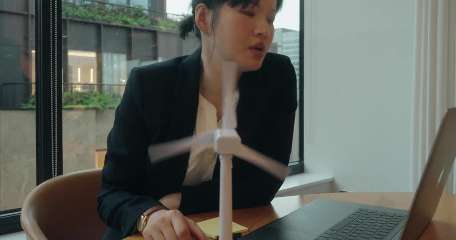 A woman in a dark blazer and white shirt is seated at a table near a window writing on a yellow sticky note a small white spinning wind turbine model sits in front of her as she works on her laptop