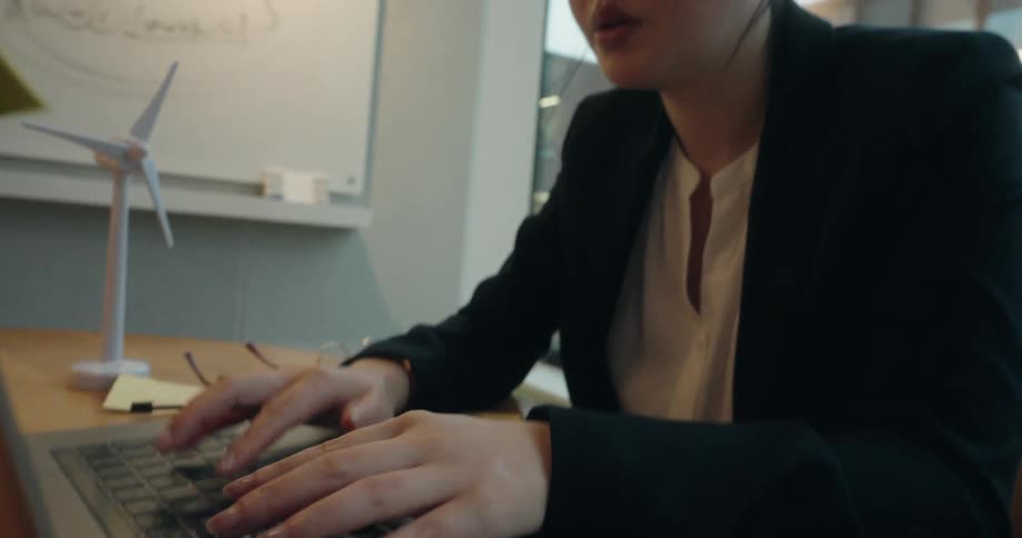 Woman sits at a table holding a model of a wind turbine and types on a laptop