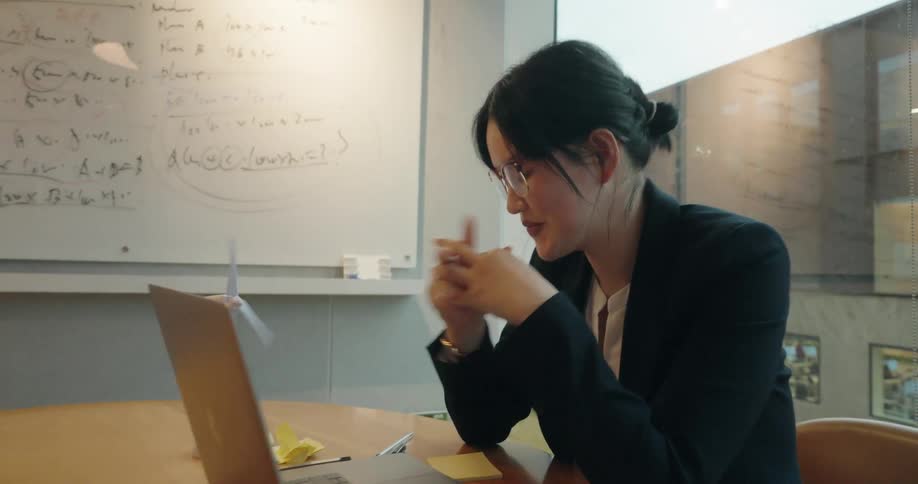 A professional woman in business attire sits at a wooden table initially looking to a laptop screen before grabbing a pen and write notes on a yellow sticky note