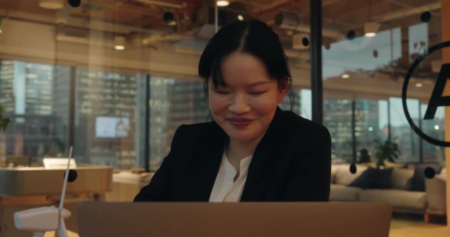 A closeup shot captures an asian businesswoman smiling and engaged in a video call on her laptop while sitting in a modern glasswalled office with city lights visible in the background