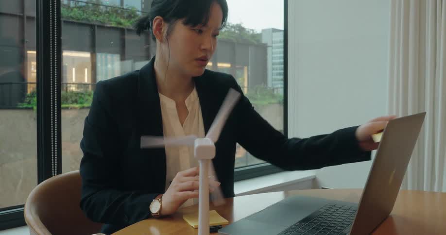 Woman in a suit looks at the laptop with a small wind turbine model spinning on the table