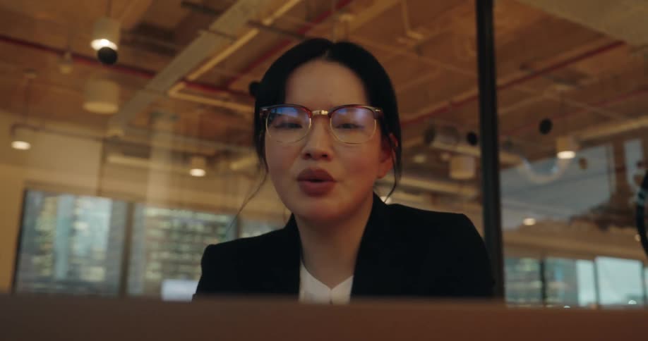 A close view of a professional woman wearing glasses and a black blazer speaking intently to a laptop camera during a video conference in a modern office space