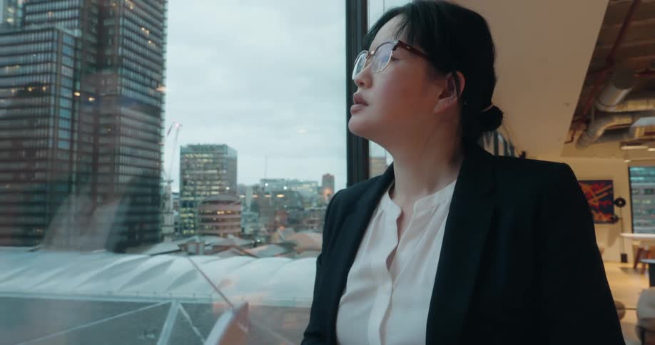 An asian businesswoman wearing glasses stands by a large office window overlooking a city skyline thoughtfully examining a small model of a wind turbine held in her hands