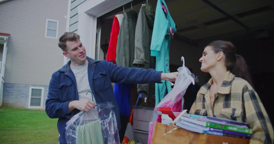 A man is standing outside a garage handing a woman a bright pink garment on a hanger while several other coats and costumes hang in the background the woman is holding a wooden crate full of books and other items as they both smile and interact