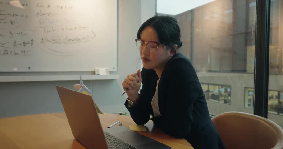 A woman looks at a sticky note while seated in an office with her laptop