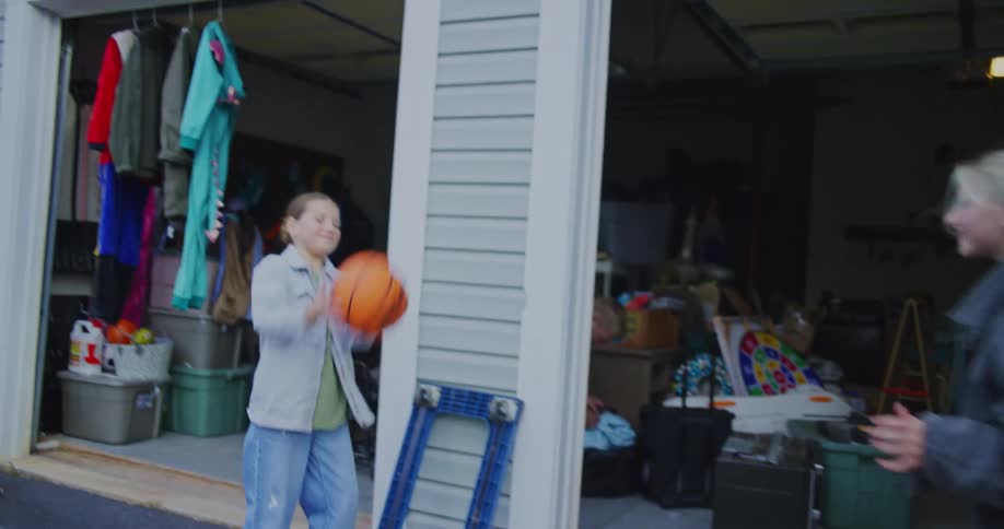 Two young girls in casual clothing are standing outside a cluttered garage playfully tossing an orange basketball back and forth between them