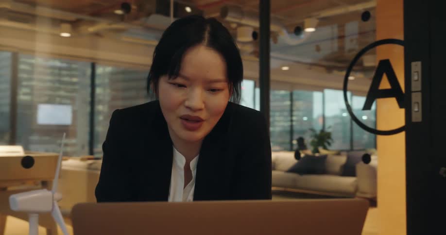 A closeup shot focuses on a young professional woman in a white shirt and black blazer intently speaking into her laptop in a modern openplan office space with city views visible through a glass wall