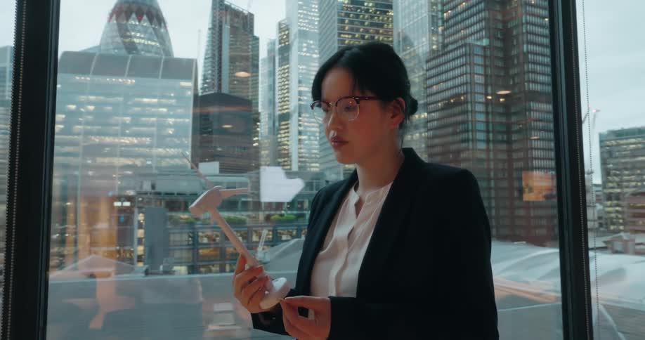 A woman wearing glasses and a business suit holds a small model of a wind turbine examining it closely while standing next to a large window with a view of a modern city skyline